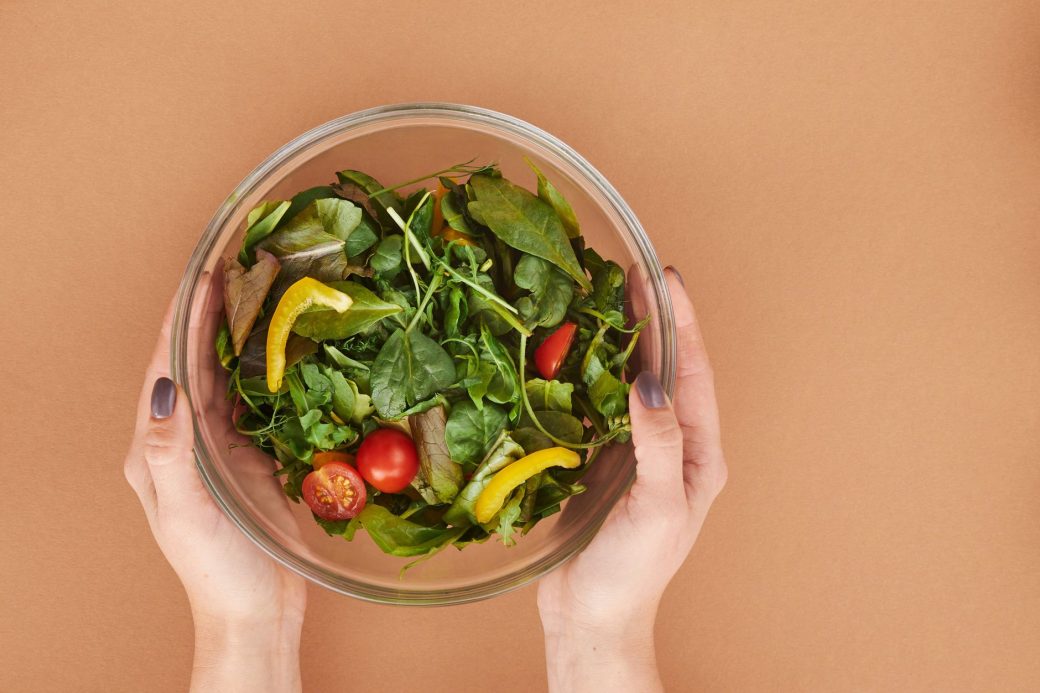 Healthy fresh salad bowl with cherry tomatoes and greens held by hands on a brown surface.