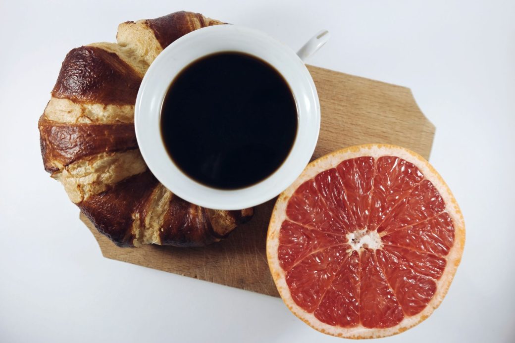 Top-down view of a coffee cup, croissant, and half a grapefruit on a cutting board.
