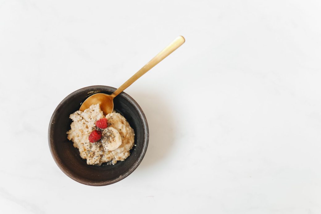 Aerial view of a breakfast bowl with oatmeal, bananas, raspberries, and chia seeds on a white surface.