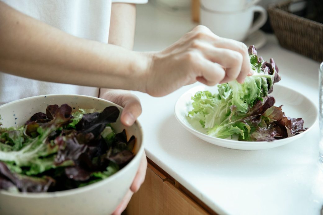 From above of crop person putting fresh mix of green salad leaves from bowl into white plate