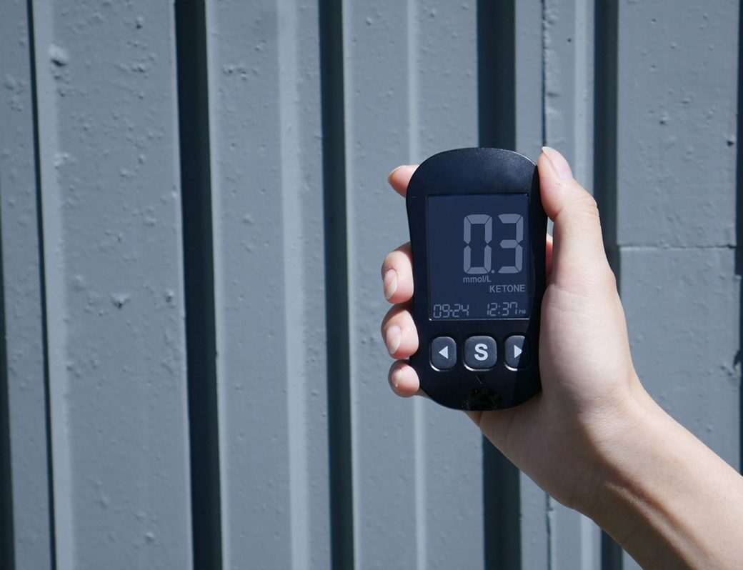 Close-up of a hand holding a ketone meter displaying ketone levels against a gray wall.