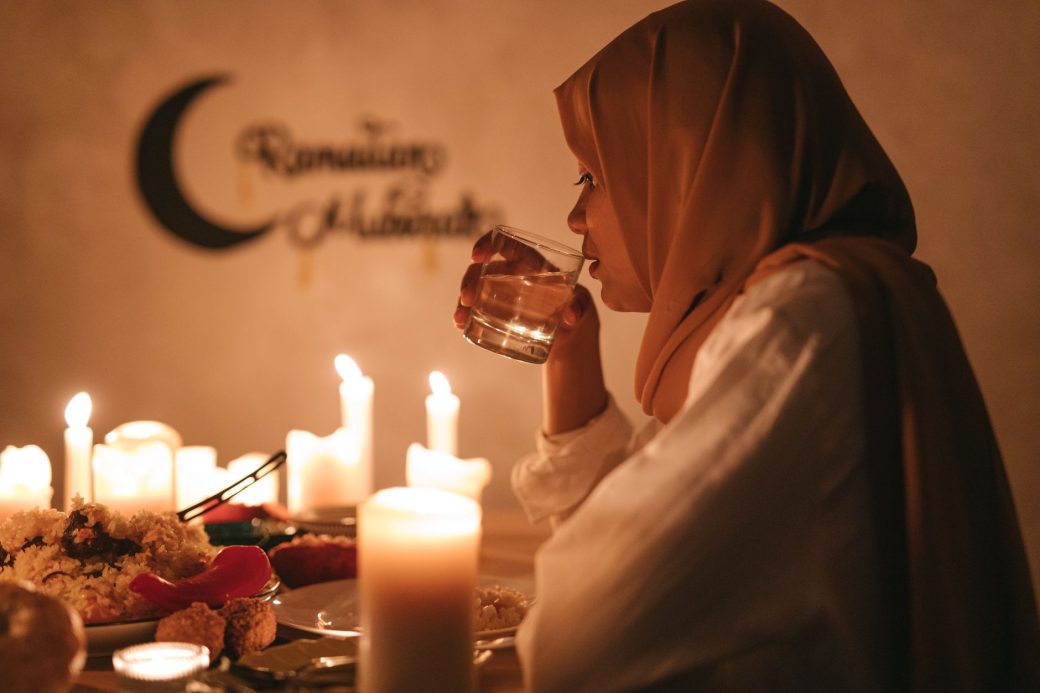 A woman in traditional attire enjoys iftar at a candlelit table during Ramadan, drinking water with a serene backdrop.