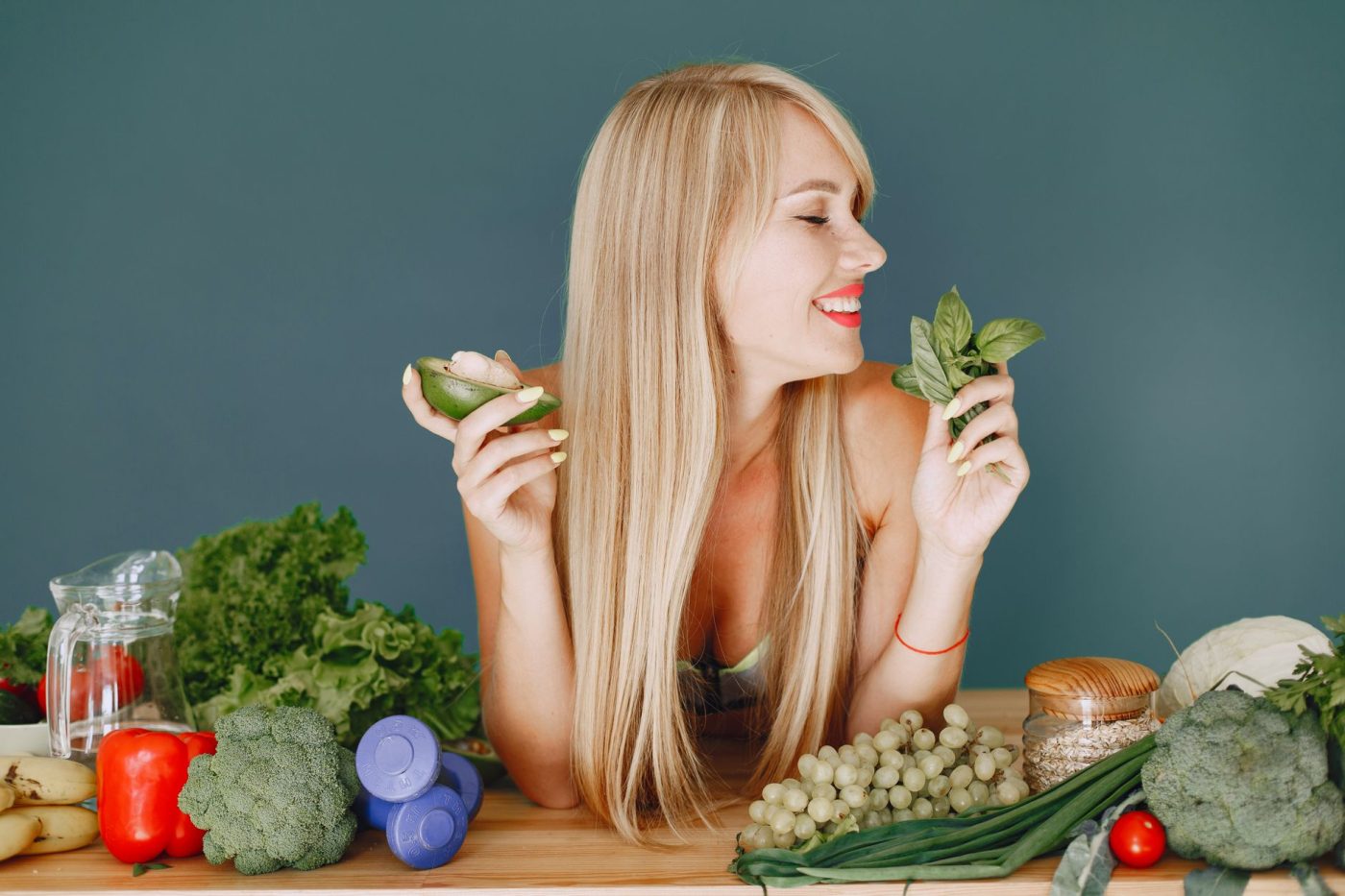 A smiling woman holding avocado and basil, surrounded by fresh vegetables and fruits for healthy eating.